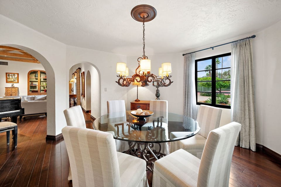 Gather around the elegant glass dining table under a striking chandelier, where warm wood floors and arched doorways create an inviting space for memorable meals with loved ones.