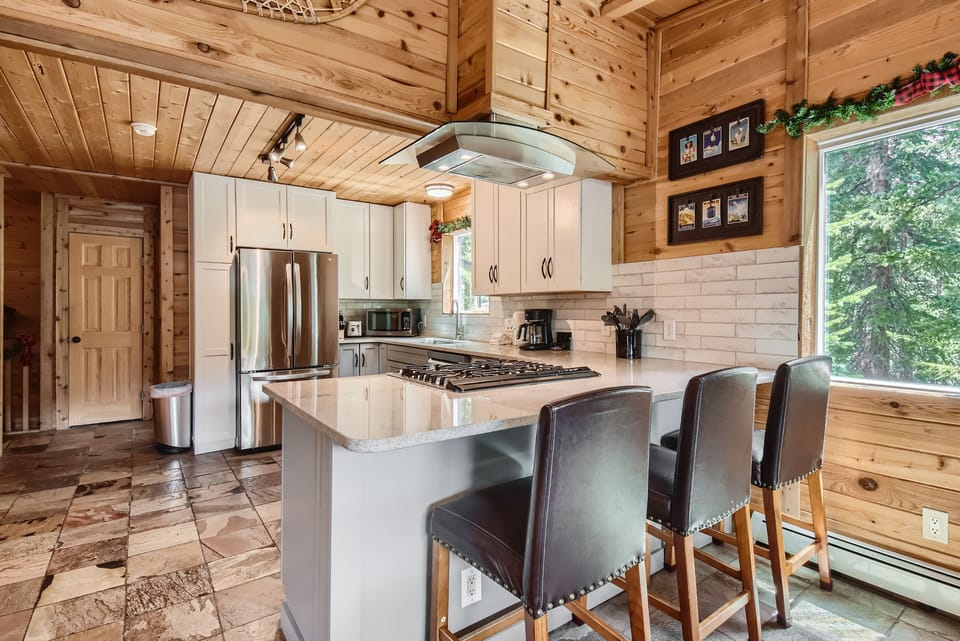 Kitchen with wooden walls and ceiling, featuring a central island with a stovetop and three black cushioned chairs. There are white cabinets, stainless steel appliances, and a large window with a view of trees.