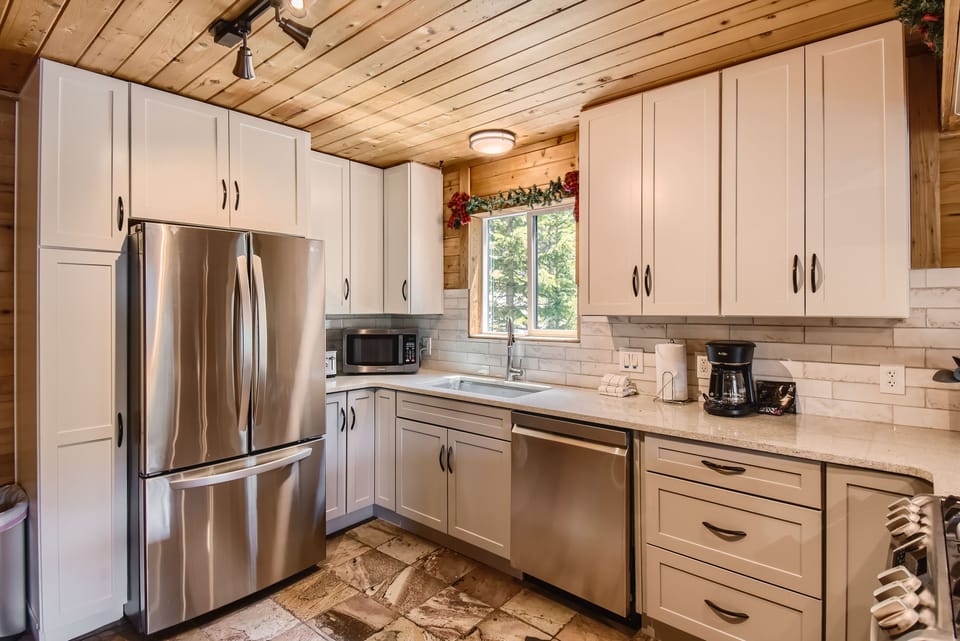 A modern kitchen with white cabinets, stainless steel refrigerator, dishwasher, microwave, and coffee maker. The walls have white subway tiles, and the ceiling is made of wood. The floor is tiled.