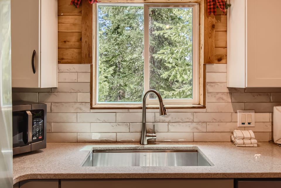 Modern kitchen sink with a stainless steel faucet, window view of forest greenery, white subway tile backsplash, white cabinets, and a microwave on the left side of the countertop.