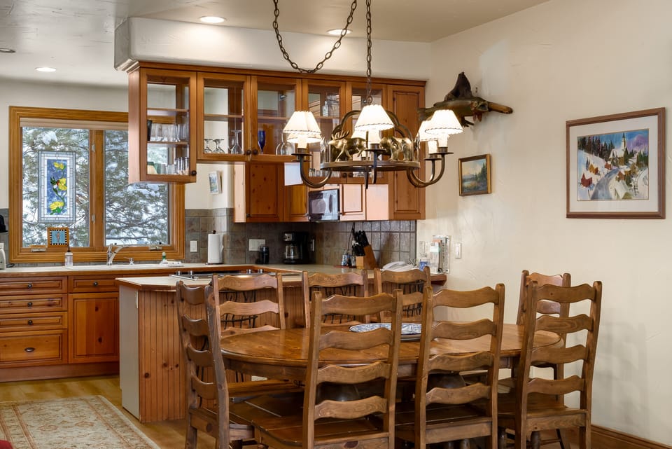 A cozy kitchen and dining area with wooden cabinets, a large wooden table with chairs, a stained-glass window, and a chandelier. Paintings and outdoor scenery are visible through the window.