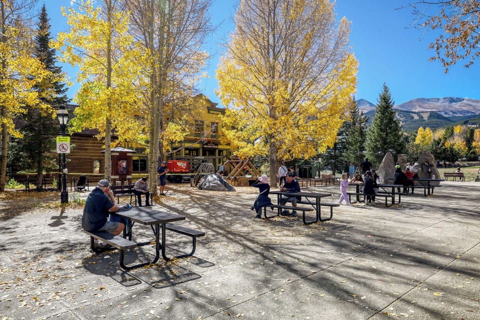 An outdoor seating area in a charming mountain town features picnic tables under brilliant golden aspens, with shops and dining nearby and majestic peaks in the distance.