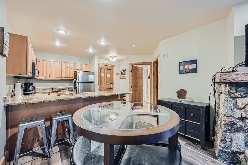 A small kitchen and dining area with wooden cabinets, stainless steel appliances, a glass-top dining table with chairs, and a breakfast bar with two stools. A decorative stone wall is visible.