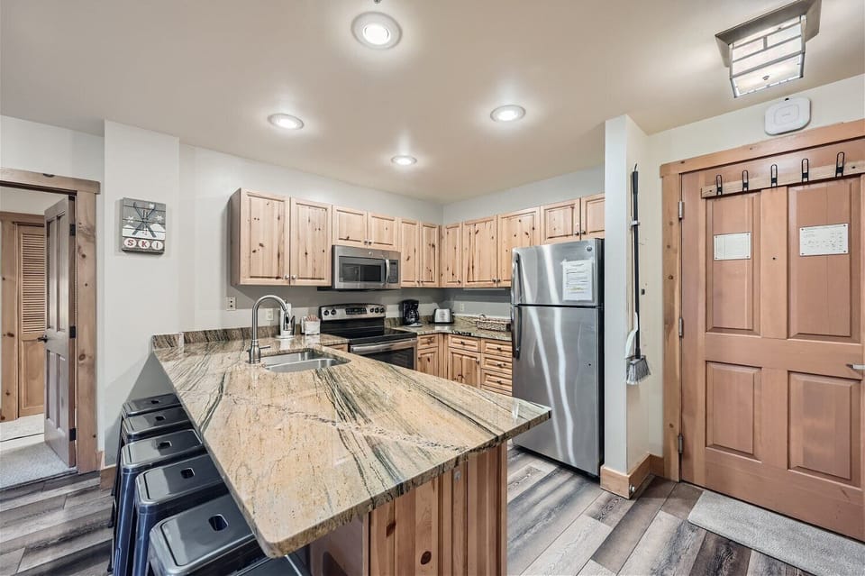 A modern kitchen with wooden cabinets, stainless steel appliances, an island with a marble countertop, and barstools. The door has notes posted on it, and the floor has a wood-like finish.