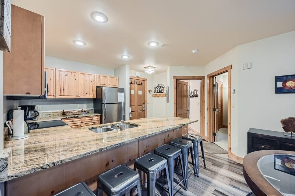 Modern kitchen with a long granite countertop, wooden cabinets, stainless steel appliances, bar stools, and recessed lighting. The background features a hallway with wooden trim and doors.