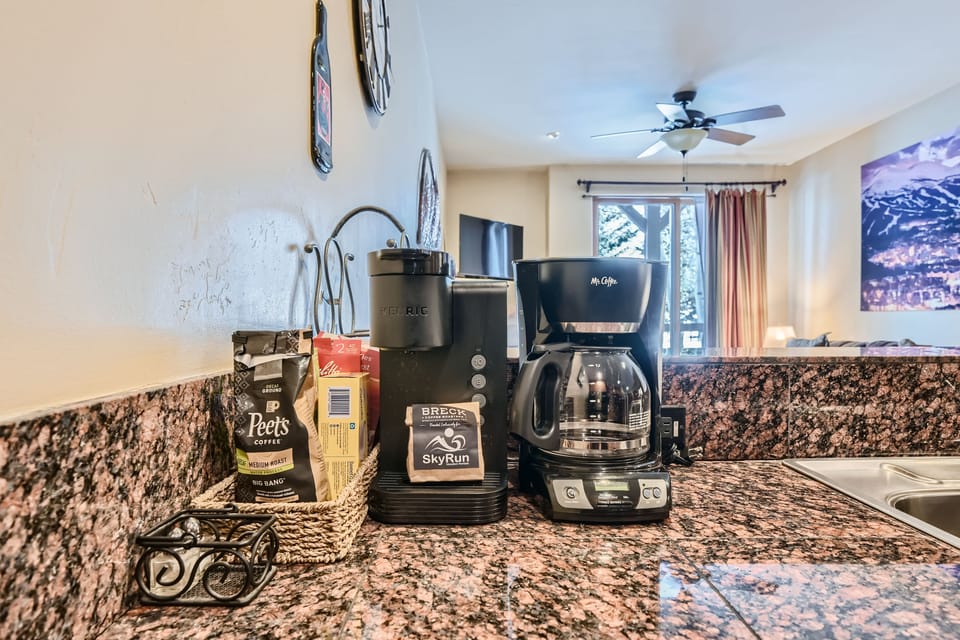 A kitchen counter with a Keurig coffee maker, a drip coffee maker, a basket with coffee supplies, and a coffee bean bag, all set on a granite surface. A window and wall art are visible in the background.