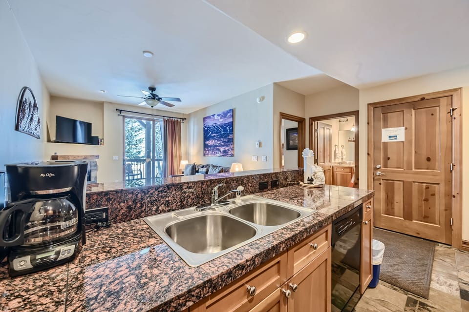 A kitchen counter with a double sink, coffee maker, and granite countertop overlooks a cozy living area with a couch, TV, ceiling fan, and large window. A bathroom is visible in the background.