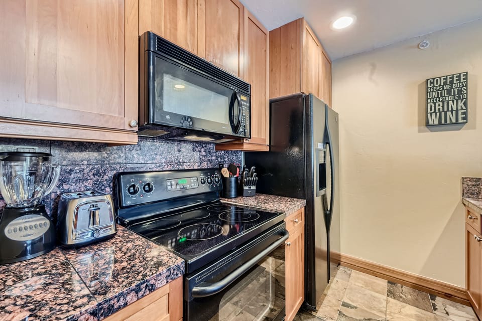 A kitchen with wooden cabinets, a black electric stove, a black microwave, a black refrigerator, a blender, and a toaster on the granite countertop. A wall sign about coffee and wine is visible.