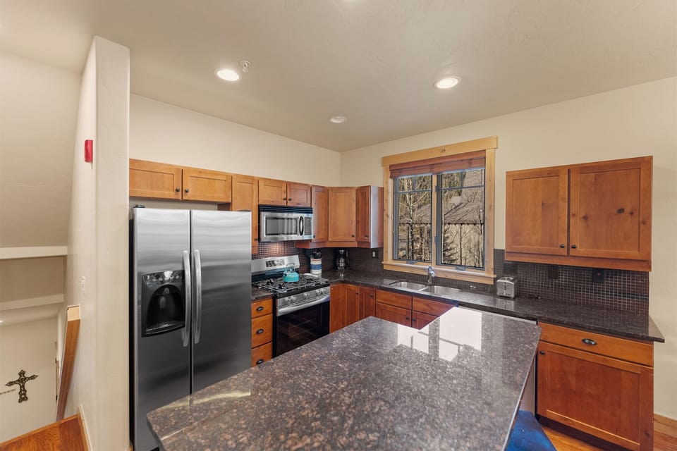 Your fully equipped kitchen features granite counters, stainless appliances, and a cozy window overlooking winter trees—perfect for preparing meals together.