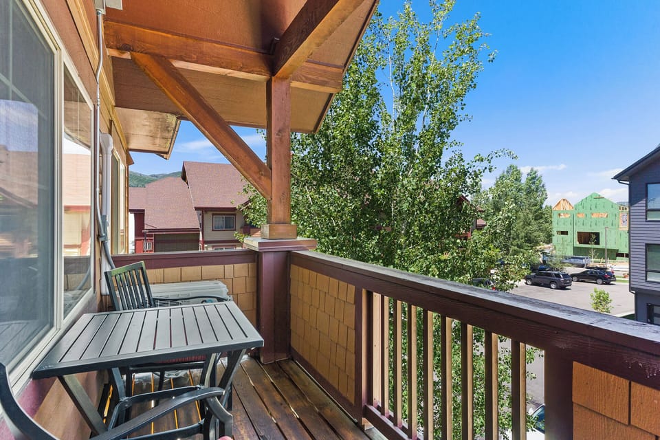 Wooden balcony with a small table and chairs overlooking a suburban neighborhood with trees and houses in the background on a sunny day.