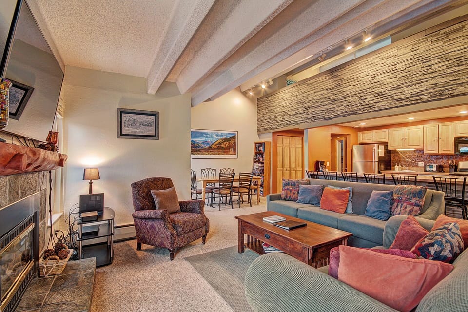 A spacious living room with a teal sectional sofa, colorful pillows, mounted TV, and coffee table. In the background, a dining area and kitchen with wooden cabinets and stainless steel appliances.