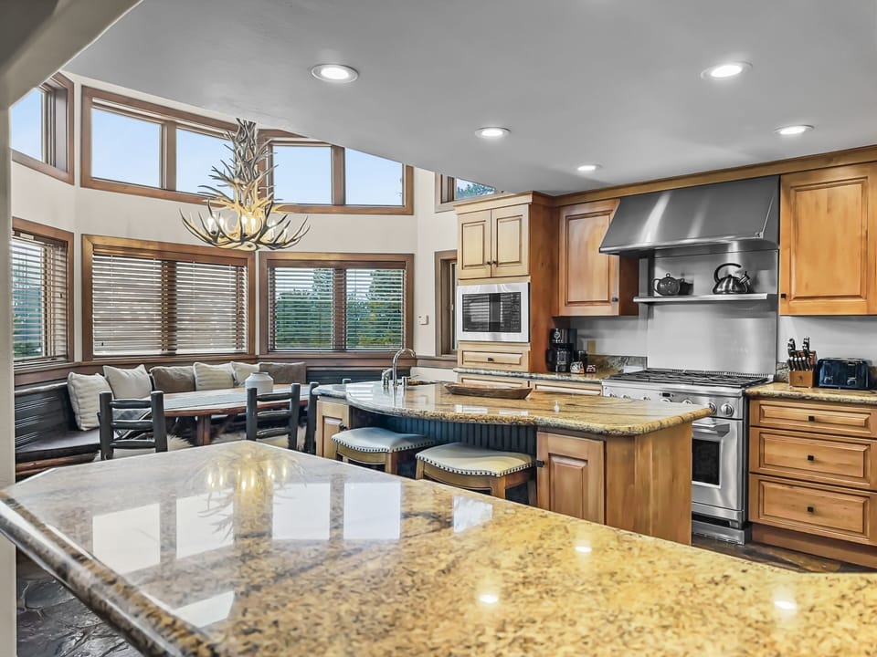 A spacious kitchen with granite countertops, a central island with seating, a gas stove, and large windows. A built-in bench sits under the windows, and a unique chandelier hangs from the ceiling.