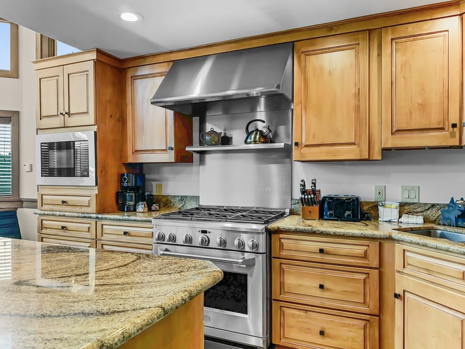 A modern kitchen with wooden cabinets, granite countertops, a stainless steel stove with a range hood, a microwave, a coffee maker, and various kitchen appliances and utensils.