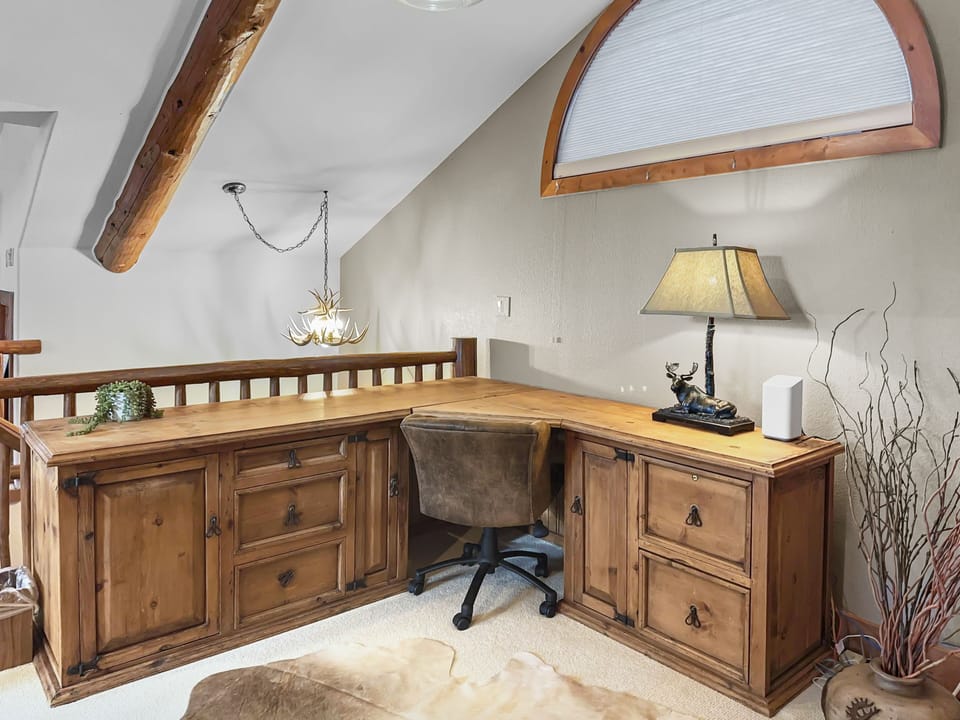 A wooden L-shaped desk with multiple drawers and cabinets is set against a wall. It includes a brown chair, a table lamp, a small plant, and a decorative antelope figurine under a large arched window.