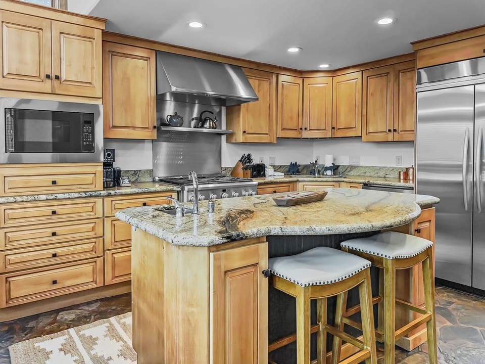 Modern kitchen with wooden cabinets, granite countertops, a central island with stools, stainless steel appliances, and a microwave. The floor is tiled, and lighting is recessed in the ceiling.