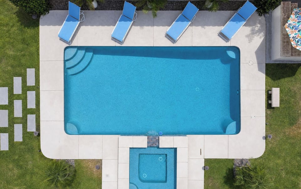 Aerial view showcasing the property's outdoor pool area, featuring a rectangular swimming pool and adjacent square hot tub, surrounded by pristine deck loungers and lush landscaping with palm trees.