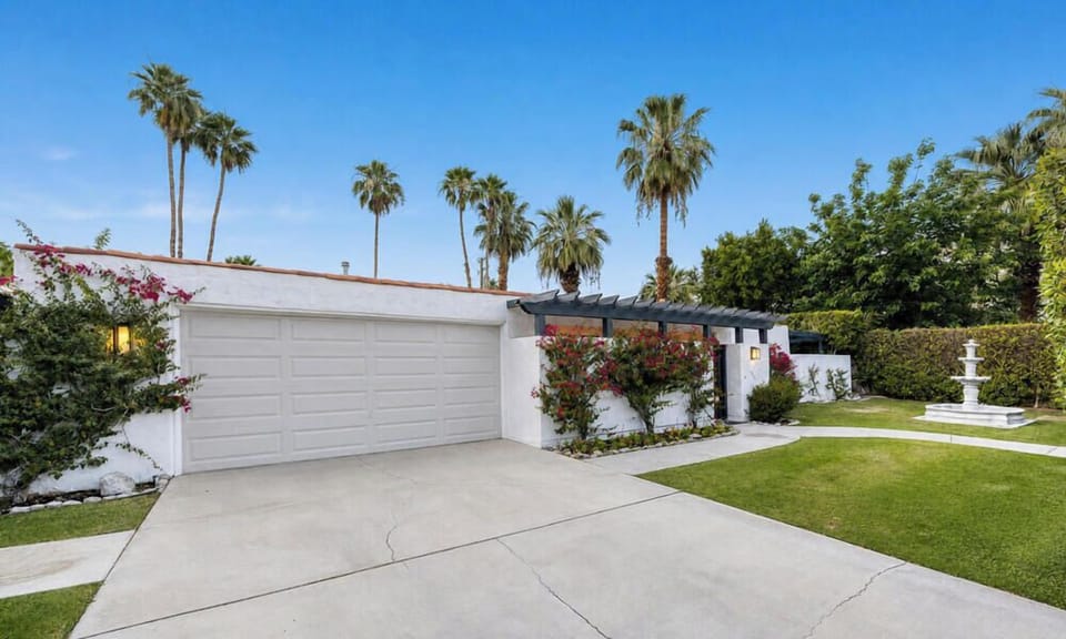 The property features a spacious driveway and lush landscaping with vibrant greenery, colorful flowering plants, and towering palm trees creating a welcoming desert oasis atmosphere.