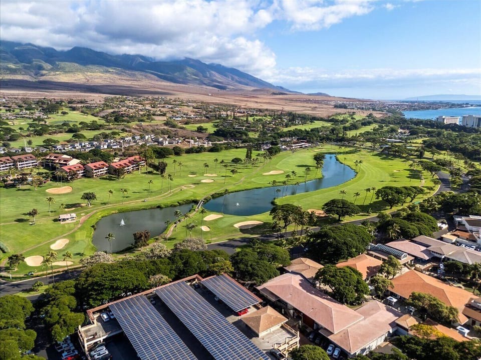Aerial view of the property nestled alongside a championship golf course, with dramatic mountain vistas and ocean glimpses defining the stunning Hawaiian landscape.