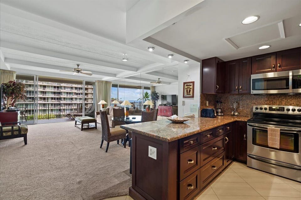 Your spacious open-plan living area features rich wood cabinetry, granite countertops, and stainless steel appliances, flowing seamlessly into a comfortable lounge and dining space with serene pool views.