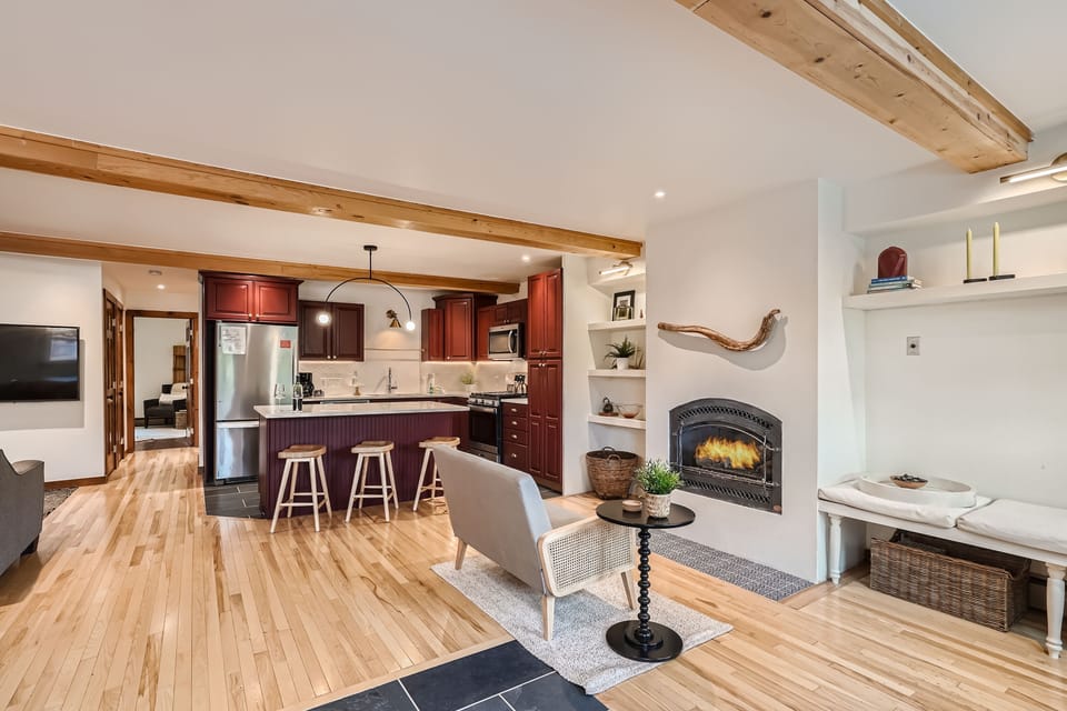 A cozy open-concept living room and kitchen with wooden flooring, beams, and cabinetry. Features a modern fireplace, bar stools at a kitchen island, and a seating area with a small table and sofa.