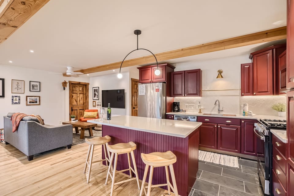 Modern kitchen and living area featuring a central island with three wooden stools, dark red cabinets, stainless steel appliances, and a gray sofa with wall art in the background.