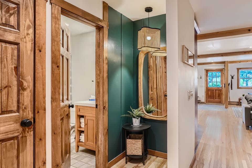 A hallway with wooden trim features a wall-mounted mirror and a small black table with a plant. An open door reveals a bathroom with a wooden vanity. A living area is visible in the background.