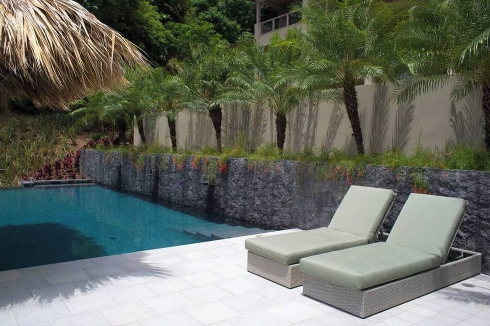 Two comfortable cushioned sun loungers situated beside a shared turquoise lap pool with natural stone waterfall walls and tropical palm trees. Relax with the best in Costa Rica luxury poolside living at this Tamarindo gated community!