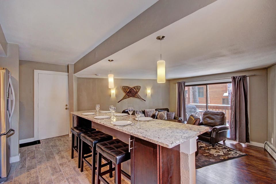 A modern kitchen with a marble island, bar stools, pendant lights, and place settings, adjoining a living area with leather sofas and large windows.