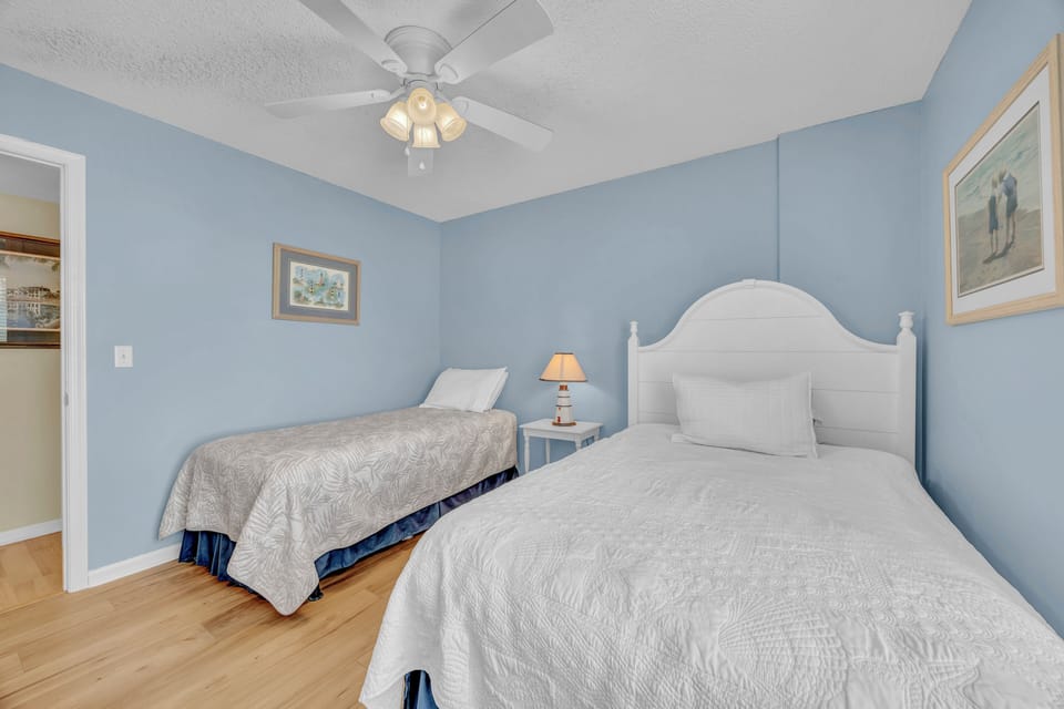 Drift off in your serene, coastal-inspired bedroom with two plush beds, soft blue walls, and a gentle ceiling fan creating the perfect ambiance for your restful retreat.