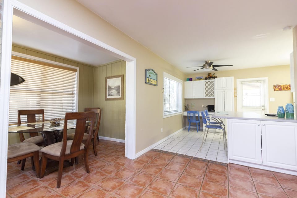 Terracotta tile flooring leads from the dining room’s wooden chairs into the white-cabinet kitchen with a ceiling fan.