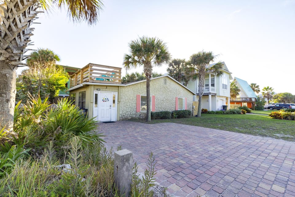 Front exterior view of the house with textured beige walls, coral shutters, and a rooftop deck surrounded by palm trees.