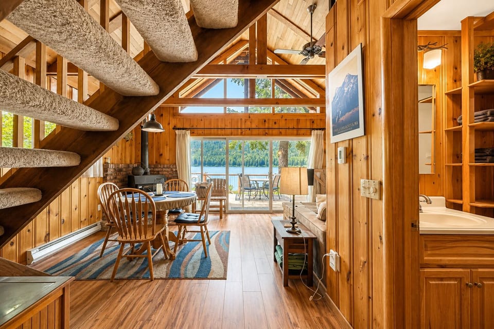 Stunning A-frame living space with vaulted ceilings, warm wood finishes, and a cozy dining area framed by breathtaking lake and mountain views through expansive windows.