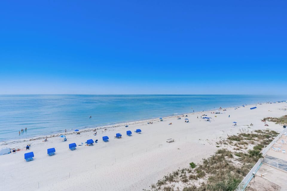 Pristine white sand beach stretches along crystal-clear waters, with blue umbrellas dotting the shoreline and gentle waves lapping the coast.