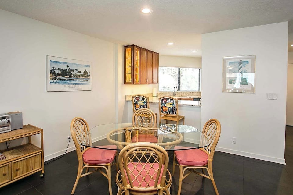 - Enjoy casual meals or board games at the glass top dining table with seating for four
- Rattan chairs with coral cushions add a tropical touch to the cozy setting
- Nearby bar seating connects the dining space to the bright kitchen