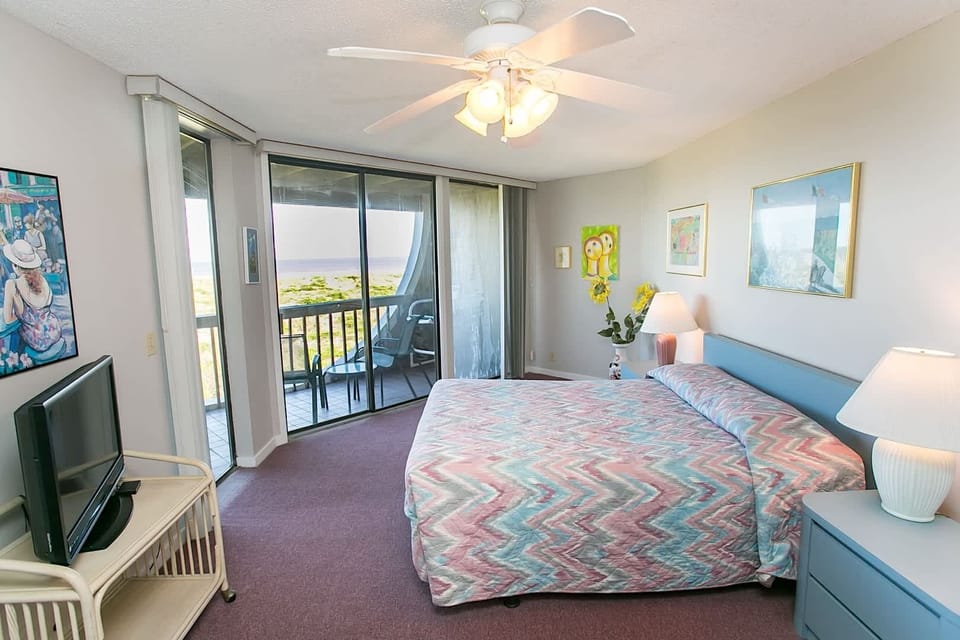- King bedroom with colorful bedding and charming coastal artwork on the walls
- Private TV for relaxed evenings, plus soft lighting and cozy decor throughout
- Sliding glass doors lead to a balcony with sweeping oceanfront scenery outside