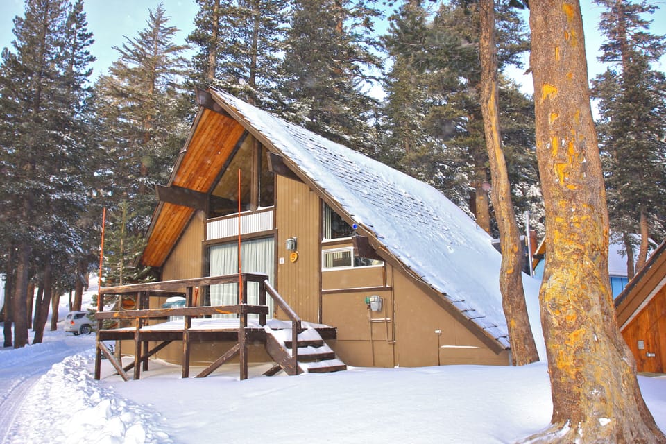 Cozy A-frame cabin nestled among towering pines, with snow-dusted deck and warm golden light filtering through winter forest.
