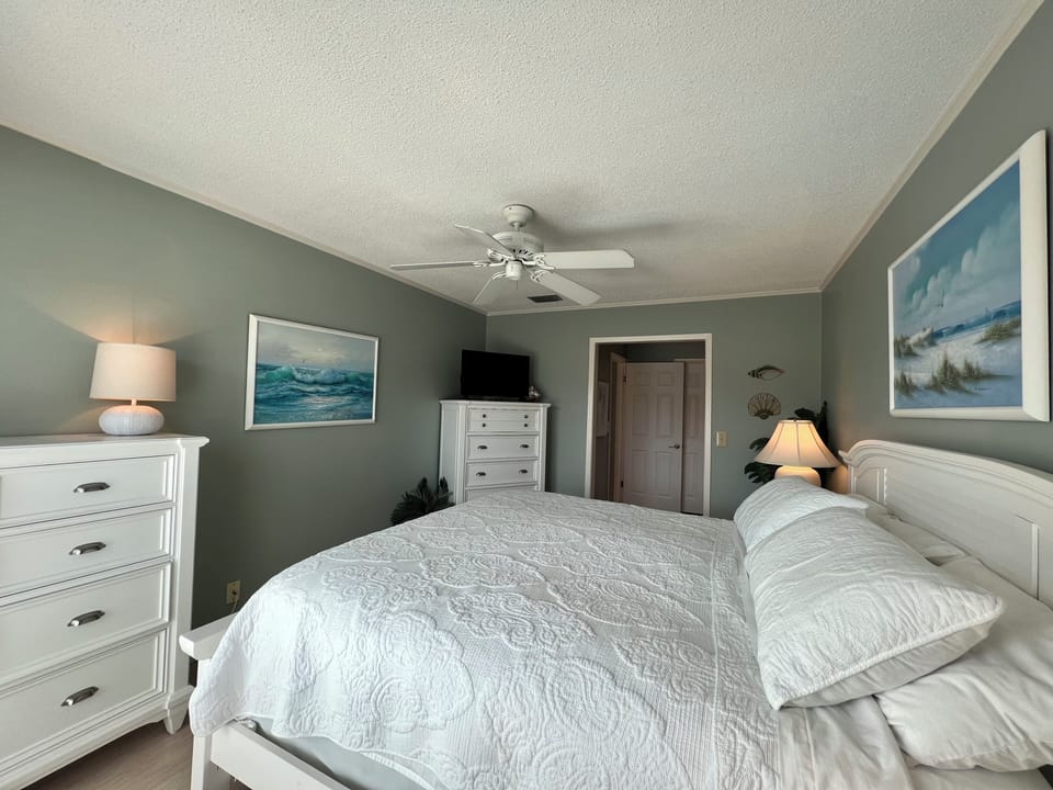 Unwind in your tranquil coastal bedroom, where soothing gray-green walls and ocean artwork create the perfect seaside sanctuary for restful nights.