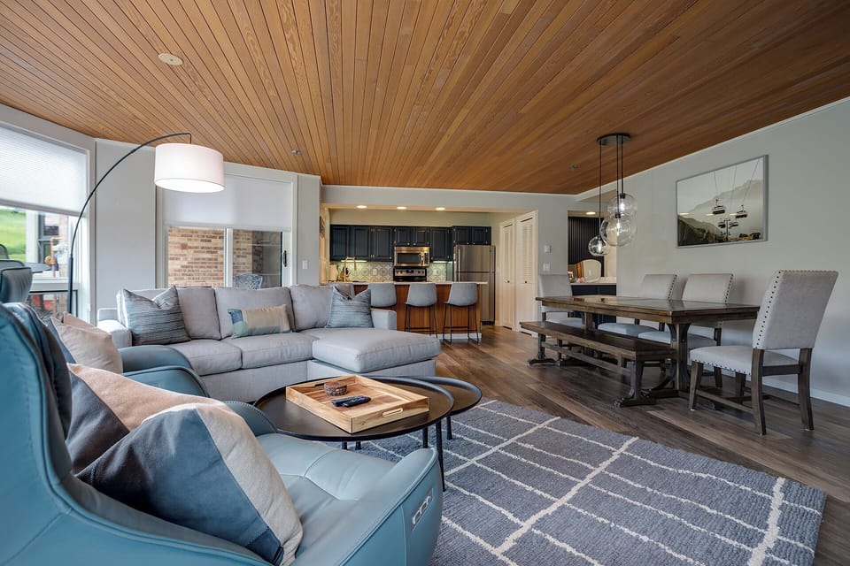 Unwind in your stylish open-concept haven featuring a plush sectional, sleek kitchen, and gorgeous wood-beam ceiling.