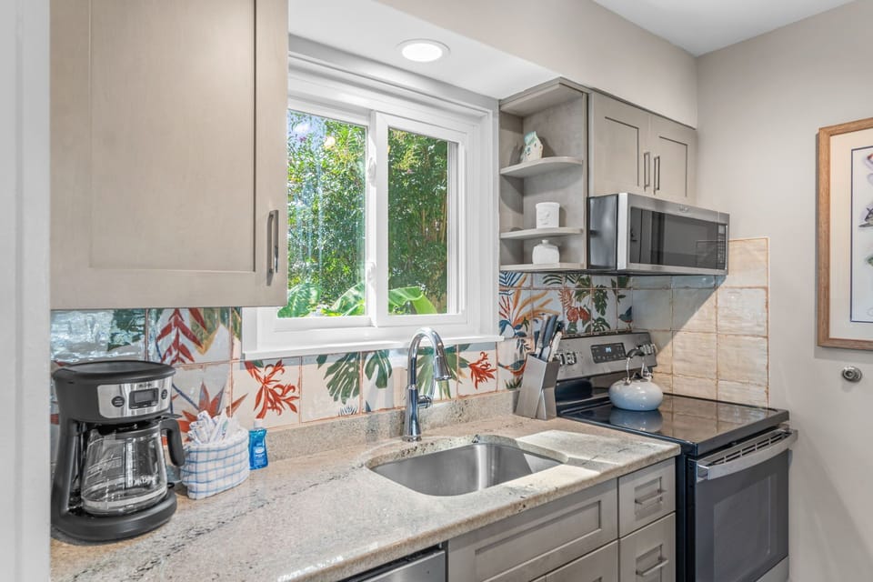 Your fully equipped kitchen awaits with colorful tile backsplash, granite counters, and modern appliances—plus a bright window framing lush greenery, making meal prep feel like a breeze.