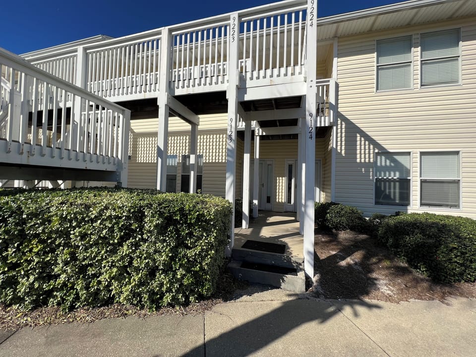 Multi-level vacation rental building featuring white railings and numbered units, set against manicured landscaping and clear blue skies.