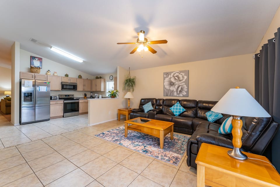 Open living area with sectional seating, coffee table, side tables, lamps, ceiling fan, and easy access to the fully equipped kitchen.