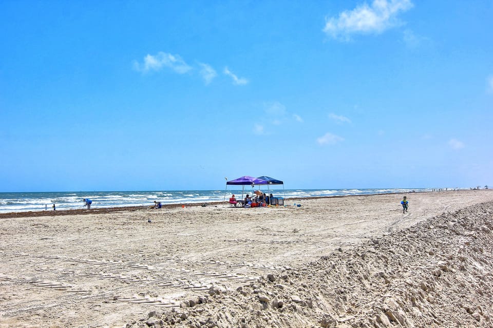 Wide sandy beach stretches along the coastline with families enjoying umbrellas and ocean waves under clear blue skies.