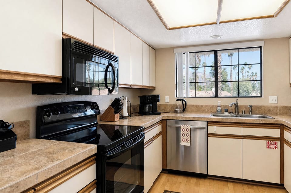 A bright, fully-equipped kitchen awaits with modern appliances, ample counter space, and natural light streaming through large windows overlooking desert palms.