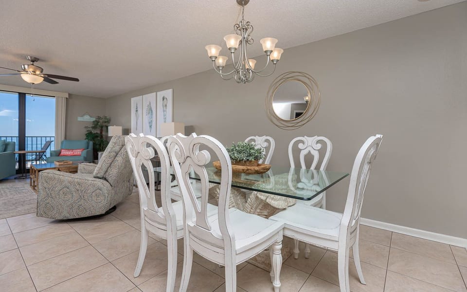Dining area features a glass table with white chairs, complemented by coastal decor and a view of the balcony