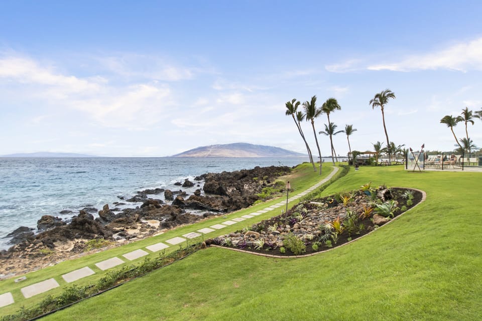 Oceanfront property with lush tropical gardens, swaying palms, and a distant island view. The rocky shoreline and manicured lawn frame a serene Hawaiian landscape.
