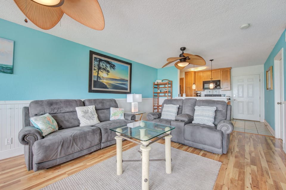 Unwind in your spacious living area featuring plush gray sectional seating, coastal-inspired décor, and an open kitchen design perfect for your beach getaway.