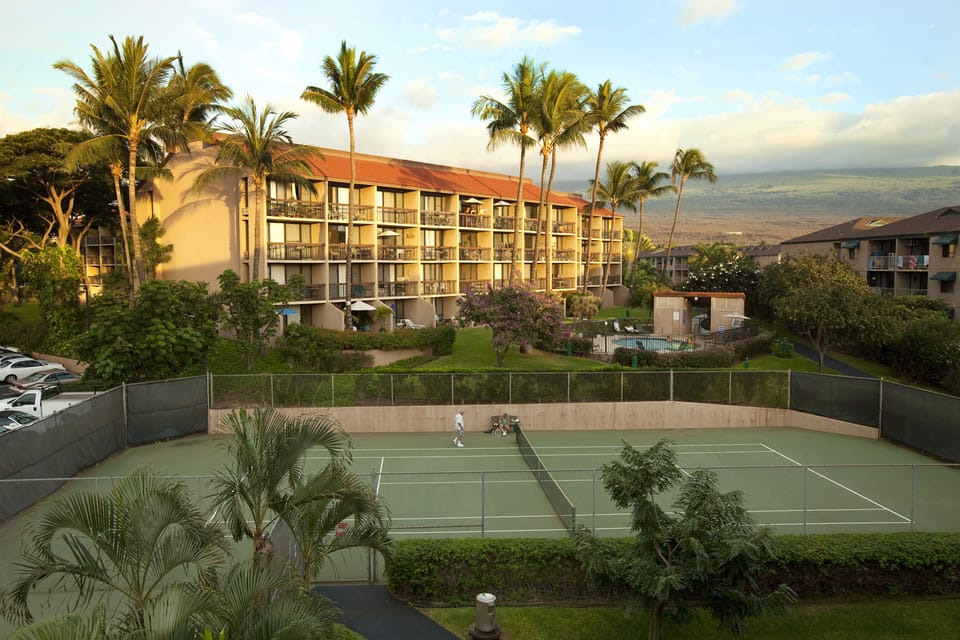 Tropical resort property featuring tennis courts, lush landscaping, and mountain backdrop. Multi-story building with balconies overlooks recreational amenities and surrounding natural scenery.