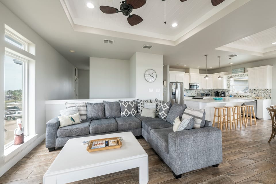 Relax in your open-concept living space featuring a plush sectional sofa, modern kitchen with waterfall countertops, and coastal ceiling fans—perfect for unwinding after a day exploring the beach.