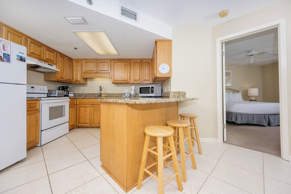 The kitchen features wooden cabinetry, a granite breakfast bar with three stools, and a view into the master suite