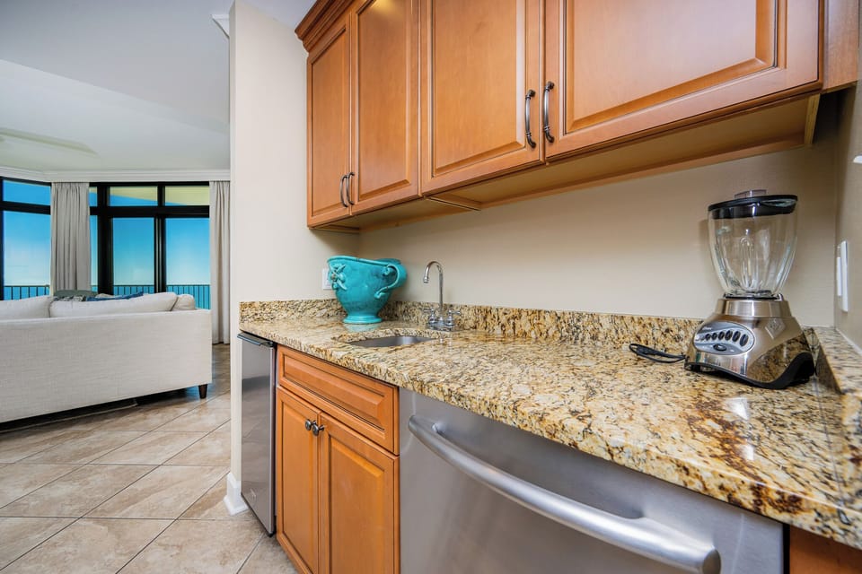 The wet bar features warm wood cabinetry, granite countertops, and a blender, with a view of the beachfront beyond